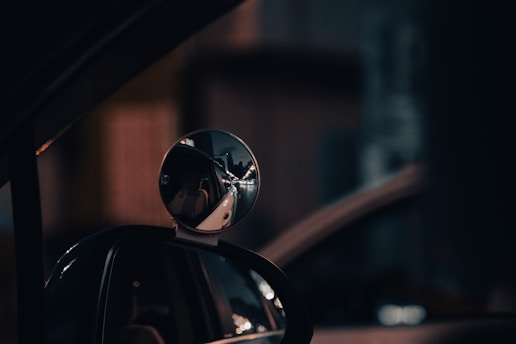A close-up view of a car's side-view mirror reflecting a dimly lit urban scene. Streetlights and a distant vehicle are visible in the reflection, creating a moody, nighttime atmosphere.
