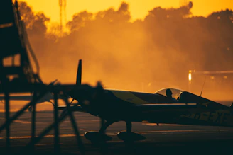 A warm golden sunrise casts long shadows on a small aircraft parked on the runway, with a pilot walking towards the cockpit, ready to take off.