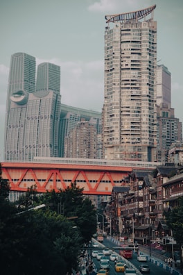 A bustling urban landscape featuring modern high-rise buildings juxtaposed with traditional architecture. An eye-catching red bridge spans across the scene, while a busy street below is filled with cars and vibrant city life.