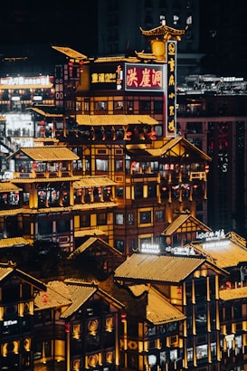 A vibrant, illuminated cityscape at night featuring traditional Chinese architectural elements. The structures are adorned with bright neon signs and lanterns, creating a lively atmosphere. The intricate design and glowing rooftops create a sense of cultural richness and modern urban life.