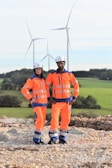 Two individuals wearing bright orange safety gear and helmets stand on a rocky surface with a backdrop of wind turbines and green fields.