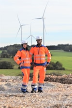 Two individuals wearing bright orange safety gear and helmets stand on a rocky surface with a backdrop of wind turbines and green fields.
