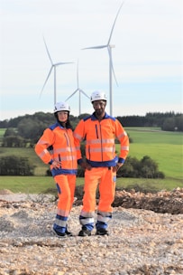 Two individuals wearing bright orange safety gear and helmets stand on a rocky surface with a backdrop of wind turbines and green fields.