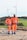Two individuals wearing bright orange safety gear and helmets stand on a rocky surface with a backdrop of wind turbines and green fields.