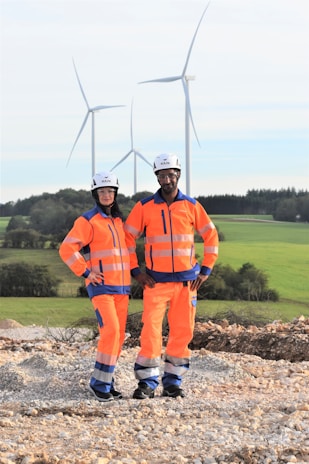 Two individuals wearing bright orange safety gear and helmets stand on a rocky surface with a backdrop of wind turbines and green fields.