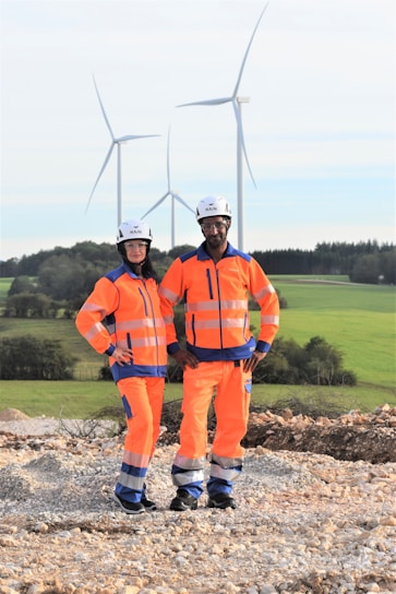 Two individuals wearing bright orange safety gear and helmets stand on a rocky surface with a backdrop of wind turbines and green fields.