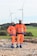 Two individuals wearing bright orange safety gear and helmets stand on a rocky surface with a backdrop of wind turbines and green fields.