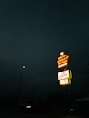 A neon-lit hotel sign stands out against the dark night sky. The sign displays 'Hotel Grand' with an advertisement for a restaurant below it. A streetlamp illuminates a nearby road where a vehicle is visible.