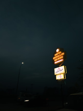 A neon-lit hotel sign stands out against the dark night sky. The sign displays 'Hotel Grand' with an advertisement for a restaurant below it. A streetlamp illuminates a nearby road where a vehicle is visible.