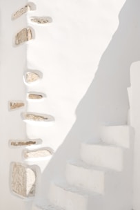 A minimalist architectural scene featuring white stone walls with embedded rocks creating a textured design. The right side shows a series of narrow, white concrete steps leading upwards, partially illuminated by sunlight casting soft shadows.