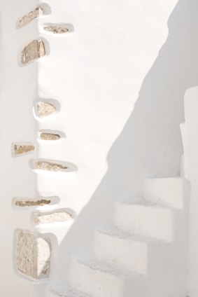 A minimalist architectural scene featuring white stone walls with embedded rocks creating a textured design. The right side shows a series of narrow, white concrete steps leading upwards, partially illuminated by sunlight casting soft shadows.