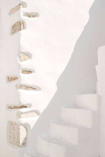 A minimalist architectural scene featuring white stone walls with embedded rocks creating a textured design. The right side shows a series of narrow, white concrete steps leading upwards, partially illuminated by sunlight casting soft shadows.