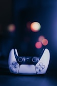 A video game controller with a sleek design is positioned centrally. The background features several out-of-focus, colorful bokeh lights creating a vibrant and slightly futuristic ambiance.