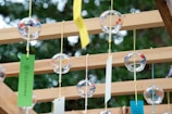 Glass wind chimes with colorful patterns are suspended from a wooden frame. Each chime has a paper strip attached, moving gently. The background shows blurred green foliage, suggesting an outdoor setting.