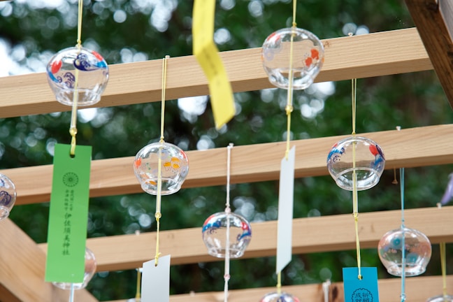 Glass wind chimes with colorful patterns are suspended from a wooden frame. Each chime has a paper strip attached, moving gently. The background shows blurred green foliage, suggesting an outdoor setting.