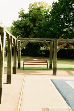 A wooden pergola structure stands over a concrete path with a blue and brown bench at its center. Lush green trees are in the background, and the surrounding area appears to be a park or garden space.