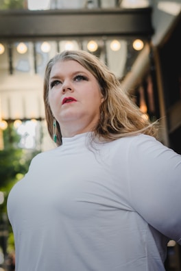 A person with long hair and light makeup is wearing a white shirt, looking upwards with a confident expression. The background includes a structure with hanging lights and green foliage.