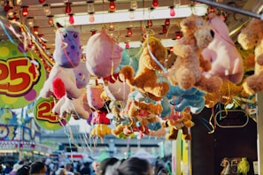 A collection of colorful plush toys, including unicorns and bears, are hanging from the ceiling of a carnival game booth. The booth is decorated with vibrant signs displaying the price of twenty-five cents. The background shows a busy fairground with people walking around and other stalls visible.