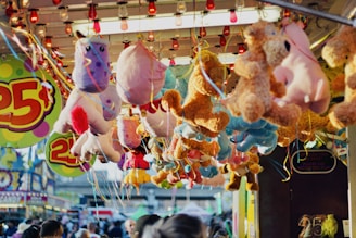 A collection of colorful plush toys, including unicorns and bears, are hanging from the ceiling of a carnival game booth. The booth is decorated with vibrant signs displaying the price of twenty-five cents. The background shows a busy fairground with people walking around and other stalls visible.