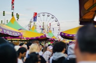 A vibrant fairground scene with a large Ferris wheel in the background. Colorful tents with yellow, green, and purple tops line the area, surrounded by a crowd of people enjoying the fair. The sky is clear, adding to the cheerful atmosphere.