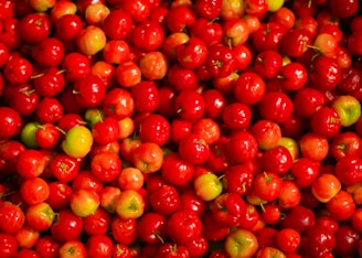 A vibrant assortment of small, red fruit densely packed together, some with hints of green. The smooth, glossy surface of the fruit reflects light, adding to the vividness of the colors.