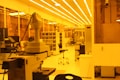 A laboratory setting with a focus on a large microscope in the foreground. The room is lit with yellow lighting, giving the space a warm, monochromatic glow. Lab equipment and furniture such as chairs and shelves are visible in the background, indicating a research or industrial work environment.