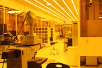 A laboratory setting with a focus on a large microscope in the foreground. The room is lit with yellow lighting, giving the space a warm, monochromatic glow. Lab equipment and furniture such as chairs and shelves are visible in the background, indicating a research or industrial work environment.