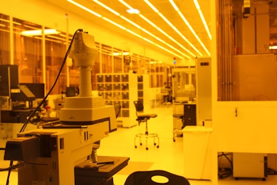 A laboratory setting with a focus on a large microscope in the foreground. The room is lit with yellow lighting, giving the space a warm, monochromatic glow. Lab equipment and furniture such as chairs and shelves are visible in the background, indicating a research or industrial work environment.