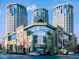 A modern shopping and office complex featuring a grand facade with large glass windows and numerous logos from various brands. The structure is flanked by high-rise towers with an architectural style combining contemporary and classic elements. Vehicles and a few pedestrians are seen on the street in front of the building.
