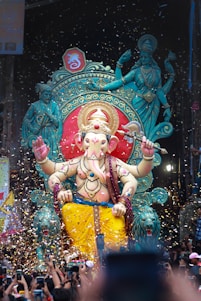 A large, colorful statue of Lord Ganesha is adorned with intricate decorations, set against a vibrant backdrop that includes ornate designs and divine figures. Confetti fills the air, adding a celebratory atmosphere. A crowd of people with raised arms and phones captures the moment, suggesting a festive event.