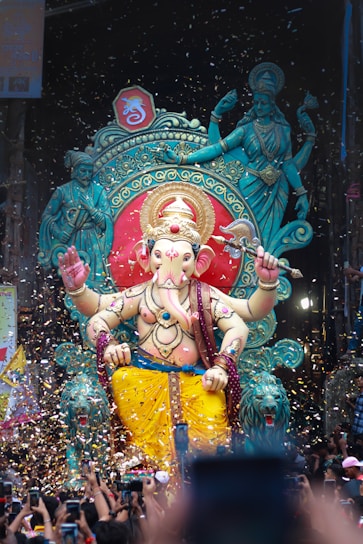 A large, colorful statue of Lord Ganesha is adorned with intricate decorations, set against a vibrant backdrop that includes ornate designs and divine figures. Confetti fills the air, adding a celebratory atmosphere. A crowd of people with raised arms and phones captures the moment, suggesting a festive event.