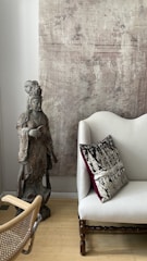 A serene interior features a wooden statue of a robed figure, standing beside a cream-colored sofa. The sofa is adorned with an elegant pillow with intricate black and cream patterns and a deep wine-red border. A textured beige wall serves as the backdrop, offering a rustic charm. The polished wooden floor is partially visible, and a woven chair is positioned in the scene.