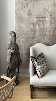 A serene interior features a wooden statue of a robed figure, standing beside a cream-colored sofa. The sofa is adorned with an elegant pillow with intricate black and cream patterns and a deep wine-red border. A textured beige wall serves as the backdrop, offering a rustic charm. The polished wooden floor is partially visible, and a woven chair is positioned in the scene.