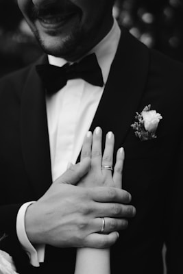 A close-up black and white photograph featuring a man in a suit with a bow tie. He is smiling, and his hands are holding and gently resting on a woman's hand, both wearing wedding rings. There is a boutonnière with a flower on his suit.