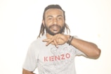 A person with a beard and dreadlocks wearing a light gray T-shirt with the word 'KENZO' in red lettering. The individual holds up two fingers in a peace sign gesture. They are wearing bracelets on one wrist, and appear relaxed against a plain white background.
