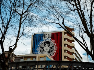 A large mural on a building features a stylized black-and-white illustration of a woman's face framed by decorative patterns and floral motifs. The words 'LIBERTE EGALITE' are boldly printed above the image. Leafless tree branches frame the sides of the scene, with a partly cloudy sky in the background.