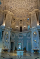 The image displays an ornate interior of a building with elaborate architectural details. The ceiling is high and intricately decorated with molded designs and circular and rectangular patterns. The walls are adorned with decorative plasterwork, statues, and arched doorways. The color scheme consists predominantly of pale blues and creams, giving a sense of elegance. A small model is placed on a pedestal in the center of the polished marble floor.