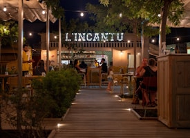An outdoor restaurant scene at night featuring multiple tables with people enjoying their meals. The ambiance is cozy and well-lit with hanging lights, and a sign displaying 'L'INCANTU' is visible in the background. There's a mix of greenery, wooden furniture, and a picturesque pathway leading into the seating area.