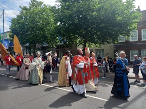 A parade features participants in historical costumes walking down a street. Characters include individuals in elaborate medieval or Renaissance attire and church robes. Spectators watch from the sidewalk, and the atmosphere is festive with some people capturing the moment on their phones. Trees and buildings line the street in the background.