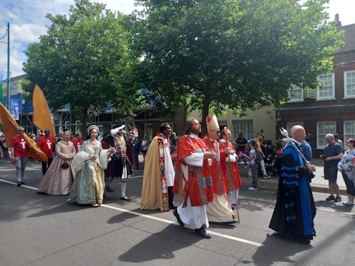 A parade features participants in historical costumes walking down a street. Characters include individuals in elaborate medieval or Renaissance attire and church robes. Spectators watch from the sidewalk, and the atmosphere is festive with some people capturing the moment on their phones. Trees and buildings line the street in the background.