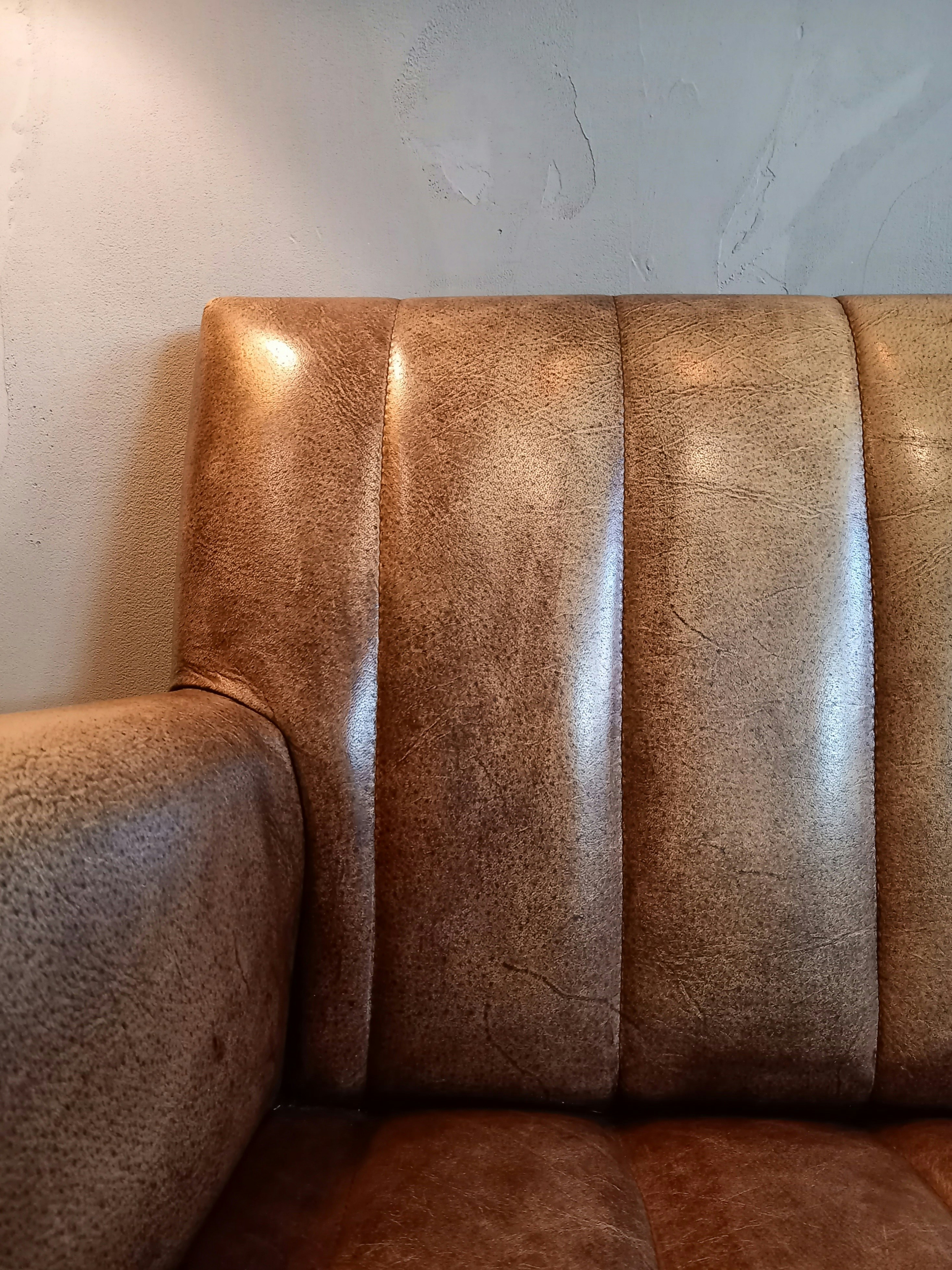 A close-up of a textured leather couch with vertical panels and a slightly worn appearance, set against a light-colored wall with subtle texture.