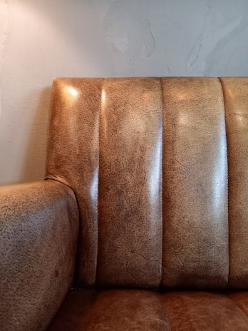 A close-up of a textured leather couch with vertical panels and a slightly worn appearance, set against a light-colored wall with subtle texture.