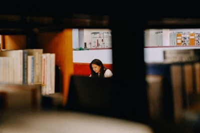 A person is seen sitting and studying in a library, surrounded by wooden shelves filled with books. The background reveals other sections of the library with additional book collections and some indoor plants.