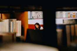 A person is seen sitting and studying in a library, surrounded by wooden shelves filled with books. The background reveals other sections of the library with additional book collections and some indoor plants.
