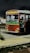 A red public bus is parked on the side of a road at night, with its headlights on and the interior lights reflecting off the windows. A green stripe runs along the side of the bus, and there are signs with writing on the front. The road is bordered by grass and a pavement with a yellow tactile paving strip, while the sky is dark and cloudy.