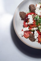 A ceramic plate contains a salad with fresh ingredients. The salad features slices of tomato, cucumber, and black olives mixed with cubes of feta cheese. Fresh herbs garnish the dish, while pieces of brown bread are placed on top for added texture.