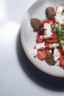 A ceramic plate contains a salad with fresh ingredients. The salad features slices of tomato, cucumber, and black olives mixed with cubes of feta cheese. Fresh herbs garnish the dish, while pieces of brown bread are placed on top for added texture.