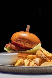 A gourmet burger with a shiny brown bun is topped with a wooden skewer. Visible inside the burger are layers of crispy fried chicken, fresh lettuce, and a slice of juicy tomato. Next to the burger, there are golden-brown potato wedges on a plate. A small white dipping sauce cup is placed near the side of the dish. The background is deep black, enhancing the vibrant colors of the food.