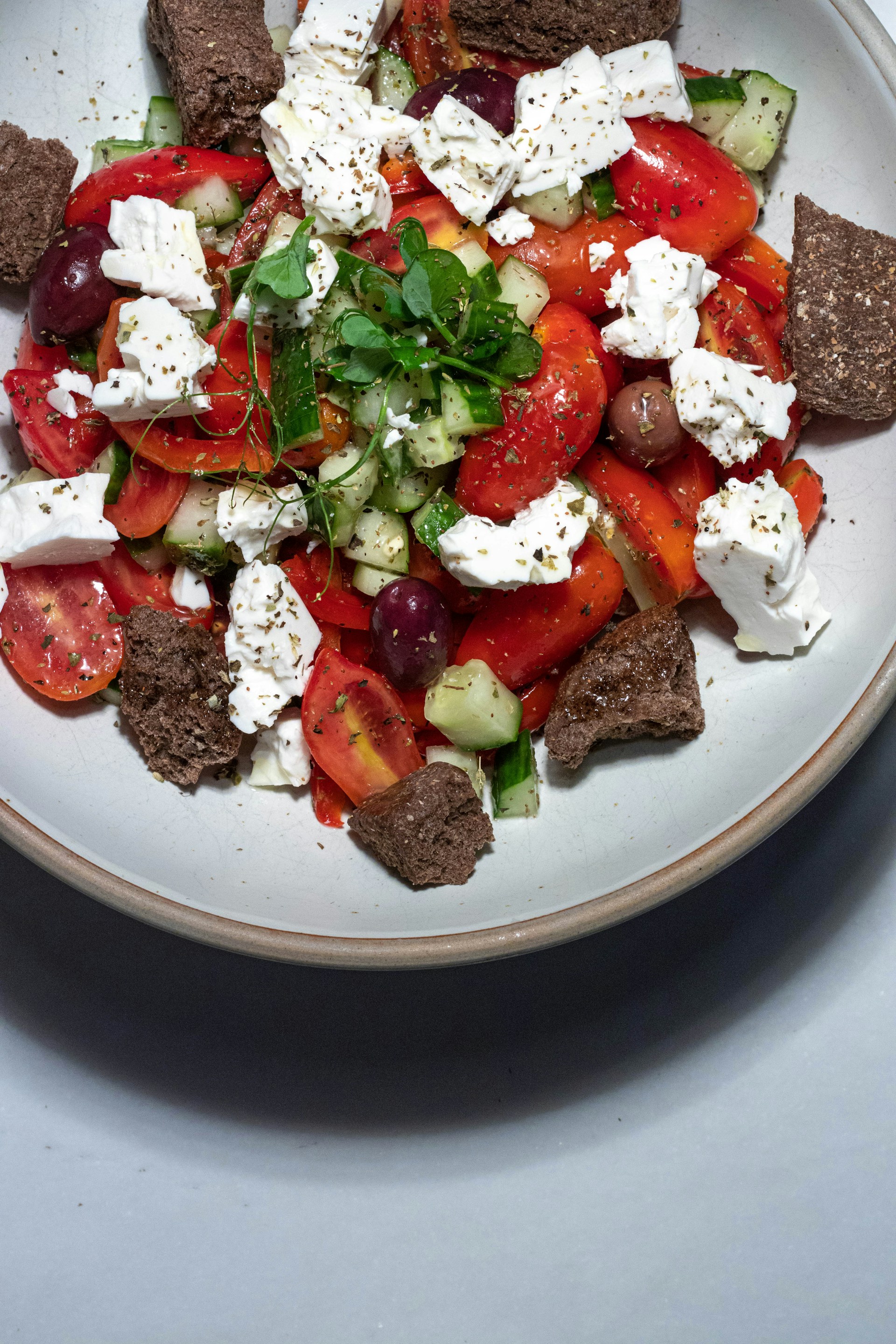 A colorful salad composed of chopped pieces of tomatoes, cucumbers, and black olives is topped with crumbled feta cheese and garnished with fresh greens. Large chunks of brown bread surround the salad on a white, round plate.