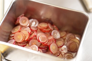 A stainless steel container filled with thinly sliced red grapes. The slices are translucent, showcasing the light and dark red hues with a hint of glossiness from the moisture. The container’s metallic finish reflects the light, adding to the overall brightness of the scene.